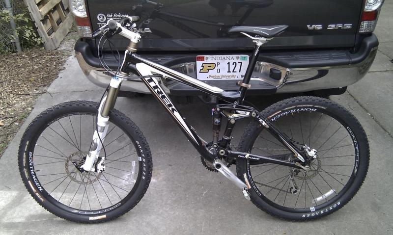 Trek Fuel EX 8: Mountain bike leaning against a vehicle, featuring a black and white frame, thick tires, and front suspension. The background includes a gravel surface and part of a pickup truck with an Indiana license plate.