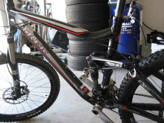 Trek Fuel EX 8: A close-up view of a Trek mountain bike, showcasing its frame, suspension system, and wheels, with a background of stacked tires and a blue container. The image captures details of the bike's components and design.