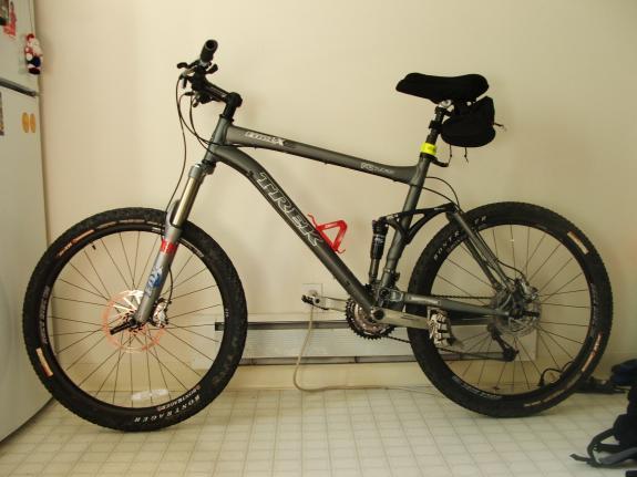 Trek Fuel EX 8: A gray Trek mountain bike with disc brakes and thick tires, positioned indoors next to a refrigerator. The bike features a black seat and visible components such as a water bottle holder and pedals. The background shows a tiled floor.