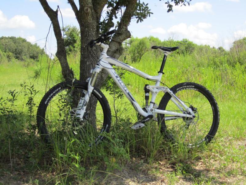 Trek Fuel EX 8: A mountain bike leaning against a tree in a green, grassy field under a blue sky with fluffy white clouds.