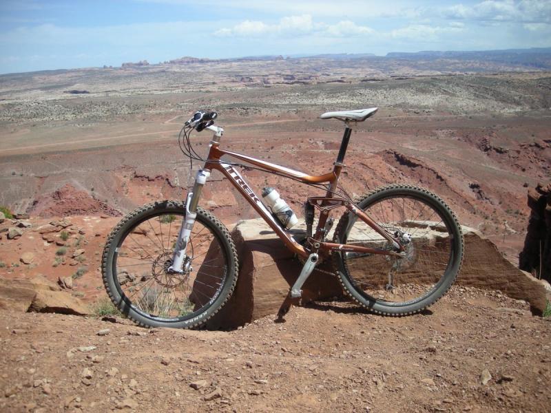 Trek Fuel EX 8: A mountain bike resting on a rocky outcrop, with a vast desert landscape in the background. The scene features red rock formations and a clear blue sky, highlighting the natural beauty of the surroundings.