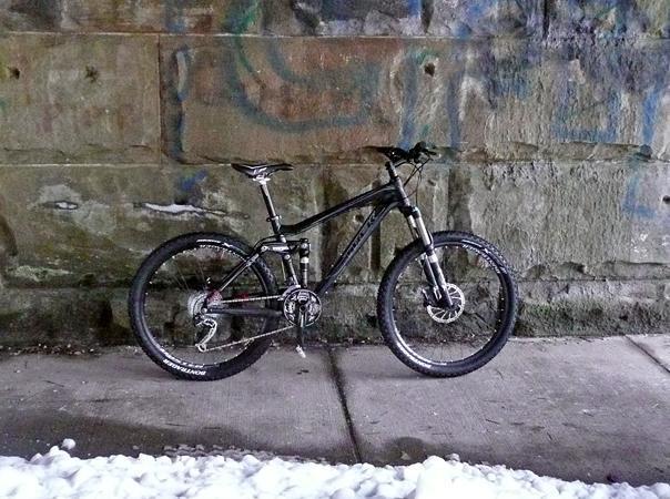Trek Fuel EX 8: A black mountain bike leaning against a textured stone wall, with a sidewalk partially covered in snow in the foreground. The bike is positioned on a concrete surface, showcasing its tires and components.