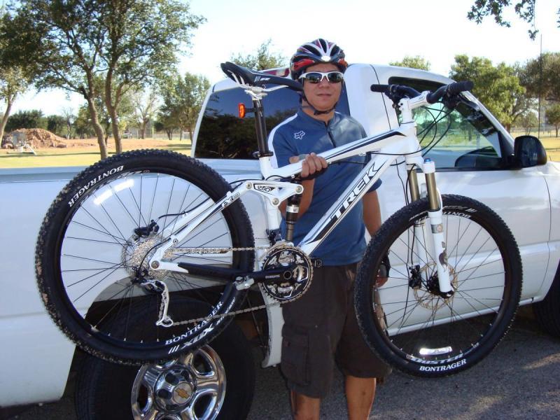 Trek Fuel EX 8: A person wearing a helmet and sunglasses is holding a white Trek mountain bike in front of a pickup truck. The background features trees and a grassy area, indicating an outdoor setting. The individual is dressed in a blue athletic shirt and shorts, showcasing their enthusiasm for cycling.