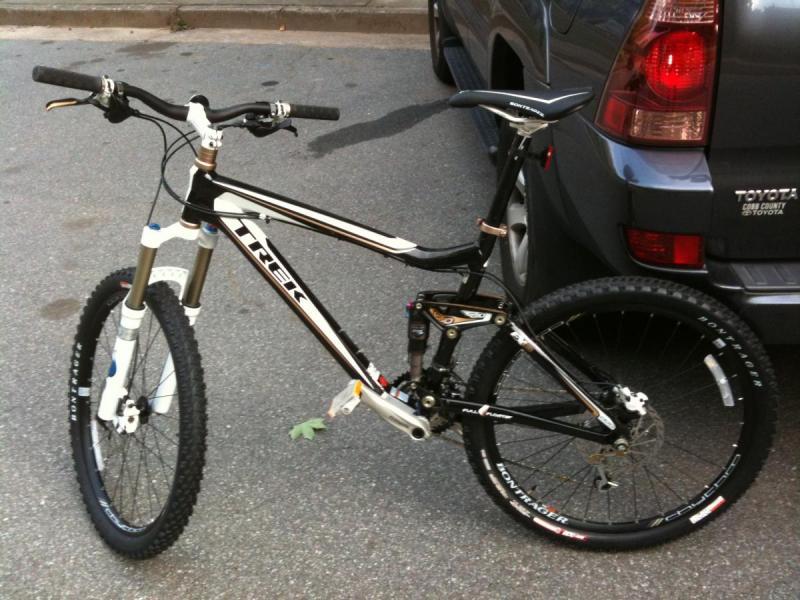 Trek Fuel EX 8: A black and white mountain bike with a sturdy frame and front suspension, parked next to a gray vehicle. The bike features prominent wheels with textured tires, and its design suggests it's built for outdoor riding. A small green leaf is resting on the bike frame.