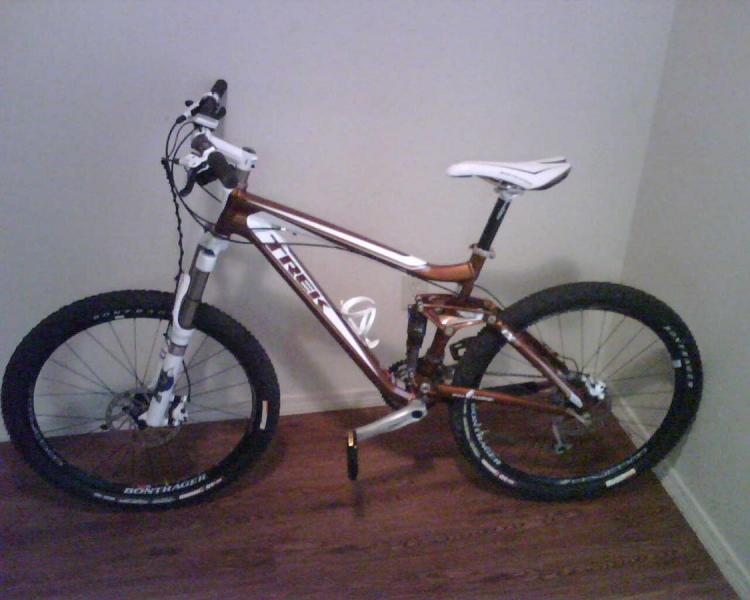 Trek Fuel EX 8: Alt tag: A mountain bike with a sleek brown and white frame, large black tires, and a white saddle, positioned against a light wall on a wooden floor.