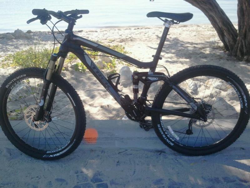Trek Fuel EX 8: A black mountain bike parked on a sandy beach, with a view of the water in the background. The bike features thick tires and a sturdy frame, positioned beside some vegetation.