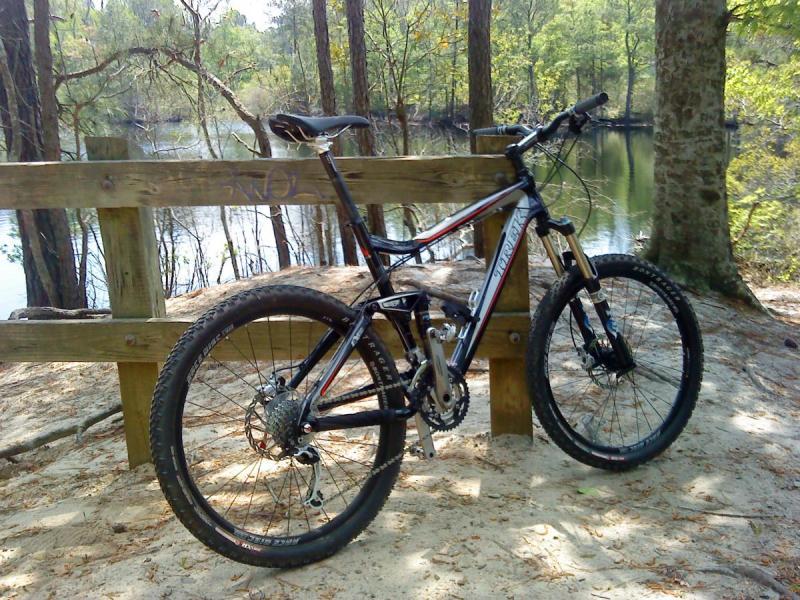 Trek Fuel EX 8: A mountain bike leaning against a wooden fence by a serene body of water, surrounded by trees. The scene captures a peaceful outdoor setting, showcasing the natural beauty of the area.