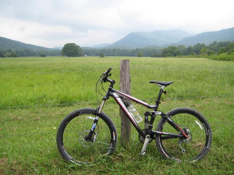 Trek Fuel EX 8: A mountain bike leaning against a wooden post, with a scenic view of a grassy field and rolling mountains in the background under a cloudy sky.