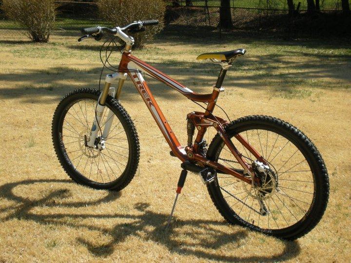 Trek Fuel EX 8: A mountain bike with a brown and silver frame, positioned on a patch of dry grass. The bike features thick tires, front suspension, and a visible gear system, with a yellow and black seat. The background includes some sparse bushes and trees, indicating an outdoor setting.