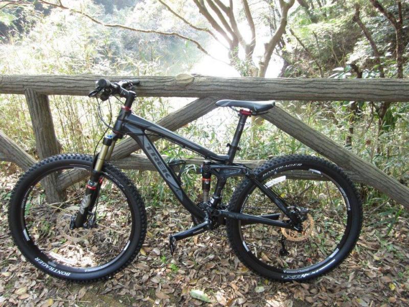 Trek Fuel EX 8: A black mountain bike is parked beside a wooden fence, surrounded by greenery and a wooded area. The sun filters through the trees in the background, creating a serene atmosphere.