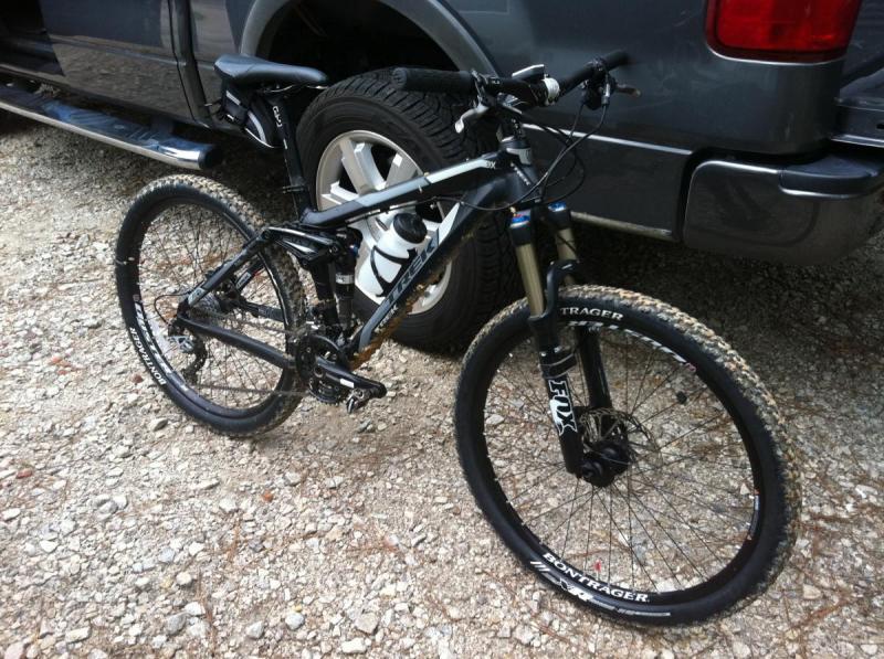 Trek Fuel EX 8: A black mountain bike parked on gravel beside a vehicle, featuring front suspension, large wheels with off-road tires, and a sleek frame design. The bike is positioned at an angle, showcasing its components, including the handlebars and seat.