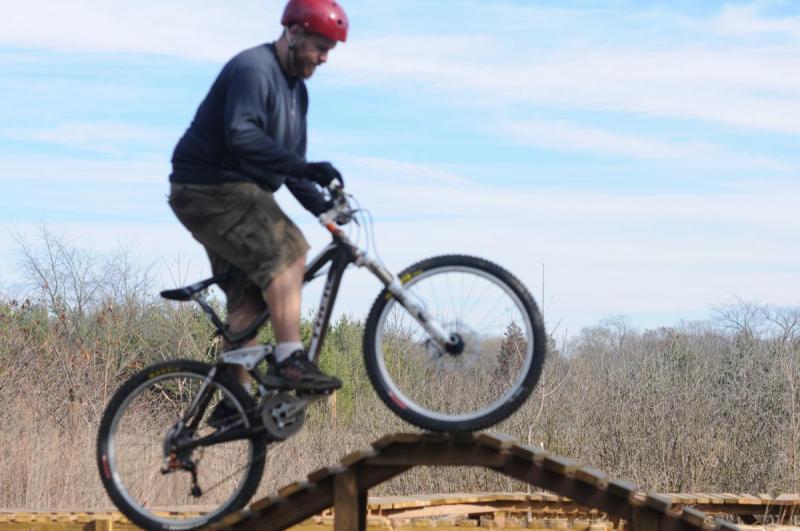 Trek Fuel EX 9: A person wearing a red helmet and gloves is riding a mountain bike over a wooden ramp in an outdoor setting. The rider is airborne, with the bike's front wheel lifted off the ground, while the background features trees and blue sky, indicating a sunny day.