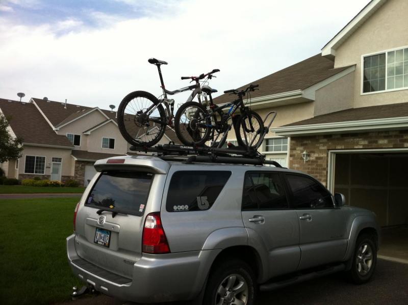 Trek Fuel EX 9: A silver SUV parked in a driveway with two mountain bikes secured on a roof rack. A residential home with a light-colored facade and green lawn is visible in the background.
