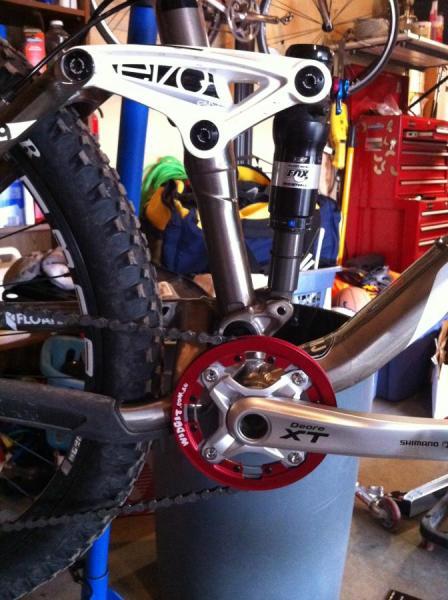 Trek Fuel EX 9: Close-up view of a mountain bike's drivetrain and suspension components. The image shows a shiny silver crankset with a red chainring, part of a Shimano XT setup, alongside a black shock absorber. The background includes various tools and bike gear in a garage setting.