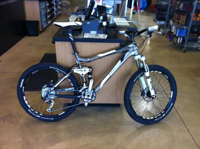 Trek Fuel EX 9: A mountain bike with a sleek gray and black frame is positioned at a retail bike shop counter. The bike features front suspension, thick tires for off-road traction, and a modern design, showcasing its suitability for rugged terrain. In the background, there are shelves filled with bike accessories and apparel.