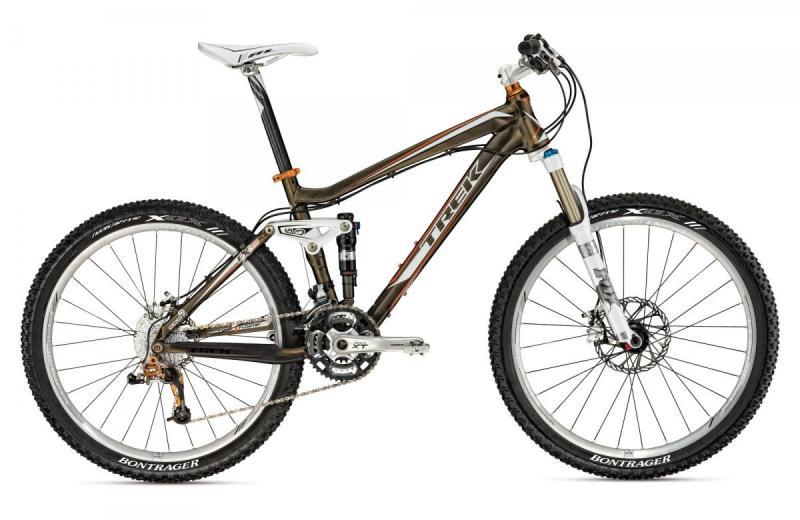 Trek Fuel EX 9: Alt text: A side view of a Trek mountain bike featuring a brown and orange frame, front suspension fork, and Bontrager wheels with knobby tires. The bicycle is positioned against a plain white background.
