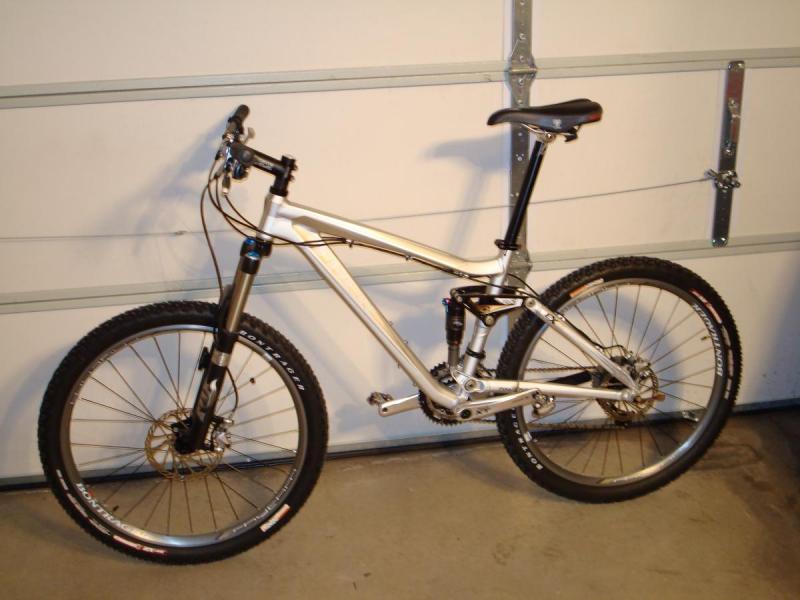 Trek Fuel EX 9: A silver mountain bike with a sleek frame and dual suspension, positioned against a garage wall with a white garage door in the background. The bike features thick tires designed for rugged terrain, disc brakes, and a black seat.