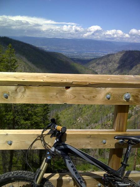 Trek Remedy 7: Mountain biking scene featuring a bike parked next to a wooden railing overlooking a scenic valley. The background includes lush green mountains and a cloudy sky, with a panoramic view of the landscape below.