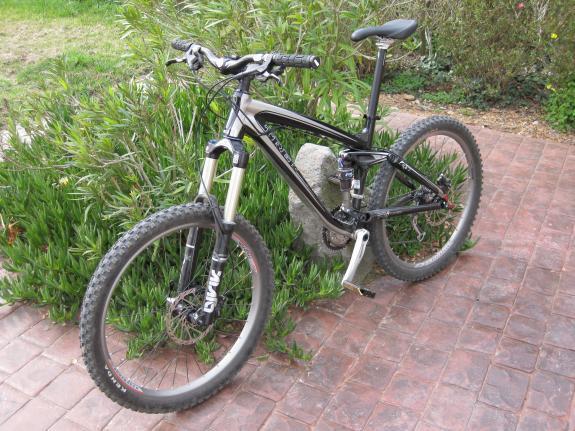 Trek Remedy 7: A black and silver mountain bike is parked on a brick pathway, surrounded by green plants. The bike features a prominent front suspension system and rugged tires, emphasizing its suitability for off-road use.
