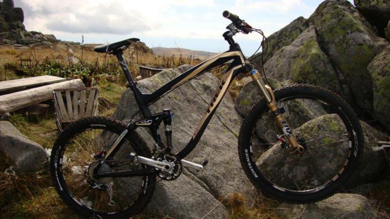 Trek Remedy 9: A mountain bike leaning against large rocks in a rugged outdoor setting, with a grassy foreground and a cloudy sky in the background. The bike features a black and gold frame, wide tires, and a suspension fork, indicating it's designed for off-road riding.