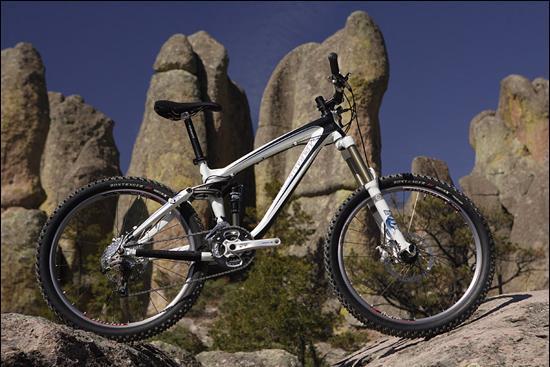 Trek Remedy 9: A mountain bike positioned on rocky terrain with large rock formations in the background under a clear blue sky. The bike features a white frame and thick tires, highlighting its suitability for rugged trails.
