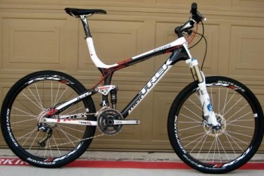 Trek Top Fuel: Alt text: A high-performance mountain bike in black, white, and red colors, featuring a lightweight frame, front suspension, and Bontrager tires. The bike is positioned on a flat surface in front of a garage door.
