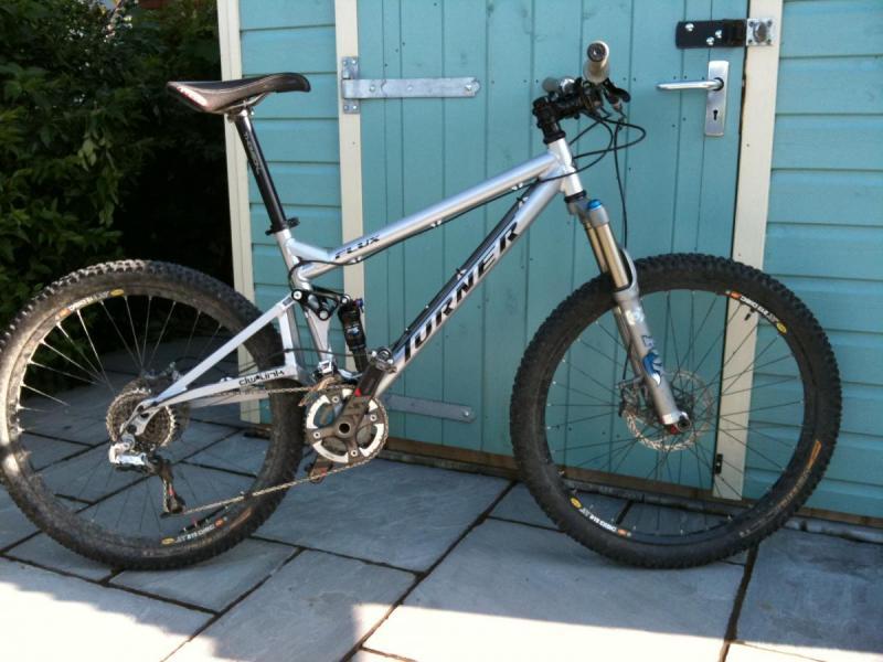 Turner Flux: A silver mountain bike leaning against a blue wooden shed, featuring large tires with tread patterns, a front suspension fork, and a visible gear mechanism. The bike is positioned on a stone patio.