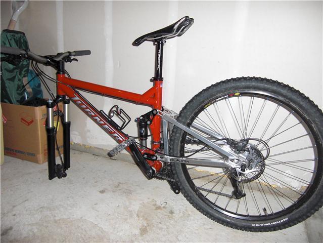 Turner Flux: A mountain bike with a bright orange frame and silver components, leaning against a wall in a garage setting. The bike features wide tires, a suspension system, and a detailed gear assembly. In the background, there are cardboard boxes and other items typical of a garage environment.