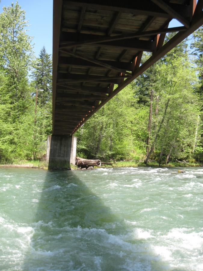 A view looking up at a wooden bridge from underneath, supported by a concrete pillar, spanning over a rushing river. The scene is surrounded by lush green trees under a clear blue sky. Sunlight creates shadows on the water's surface, enhancing the natural beauty of the setting. Larison Rock mountain bike trail.
