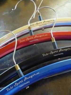 Urbane Innovations Velo Sport Hangers: A close-up image of several colorful bicycle hangers in various shades, including red, blue, and gold, each displaying the text "Velo Sport Hanger.com" and "Patent Pending." The hangers are arranged in a staggered manner against a neutral background.