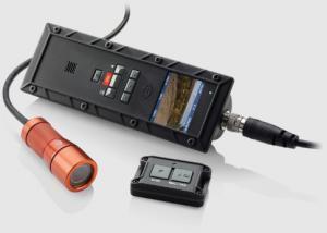 V.I.O. POV.1.5: A handheld device with buttons for controlling functions, connected to a cable and a small camera attached to a cylindrical flashlight. Beside it lies a remote control with two buttons, all set against a neutral background.