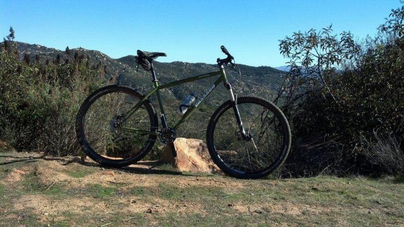Vassago Jabberwocky: A parked mountain bike on a rocky terrain with a scenic view of rolling hills and clear blue skies in the background. The bike is green and features a water bottle holder, surrounded by sparse vegetation.