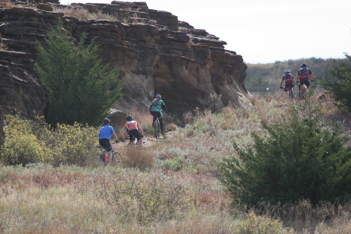 Four mountain bikers riding along a trail in a scenic landscape. The trail is surrounded by rocky cliffs and vegetation, with bushes and tall grasses visible in the foreground. The bikers are wearing helmets and colorful cycling gear, and the sky is partly cloudy. Switchgrass mountain bike trail.