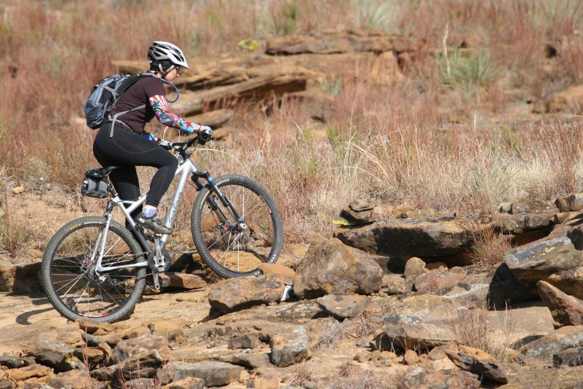 A person wearing a helmet and colorful long-sleeved shirt rides a mountain bike along a rocky terrain with sparse vegetation. The background features dry grass and scattered rocks, suggesting an outdoor biking trail. Switchgrass mountain bike trail.