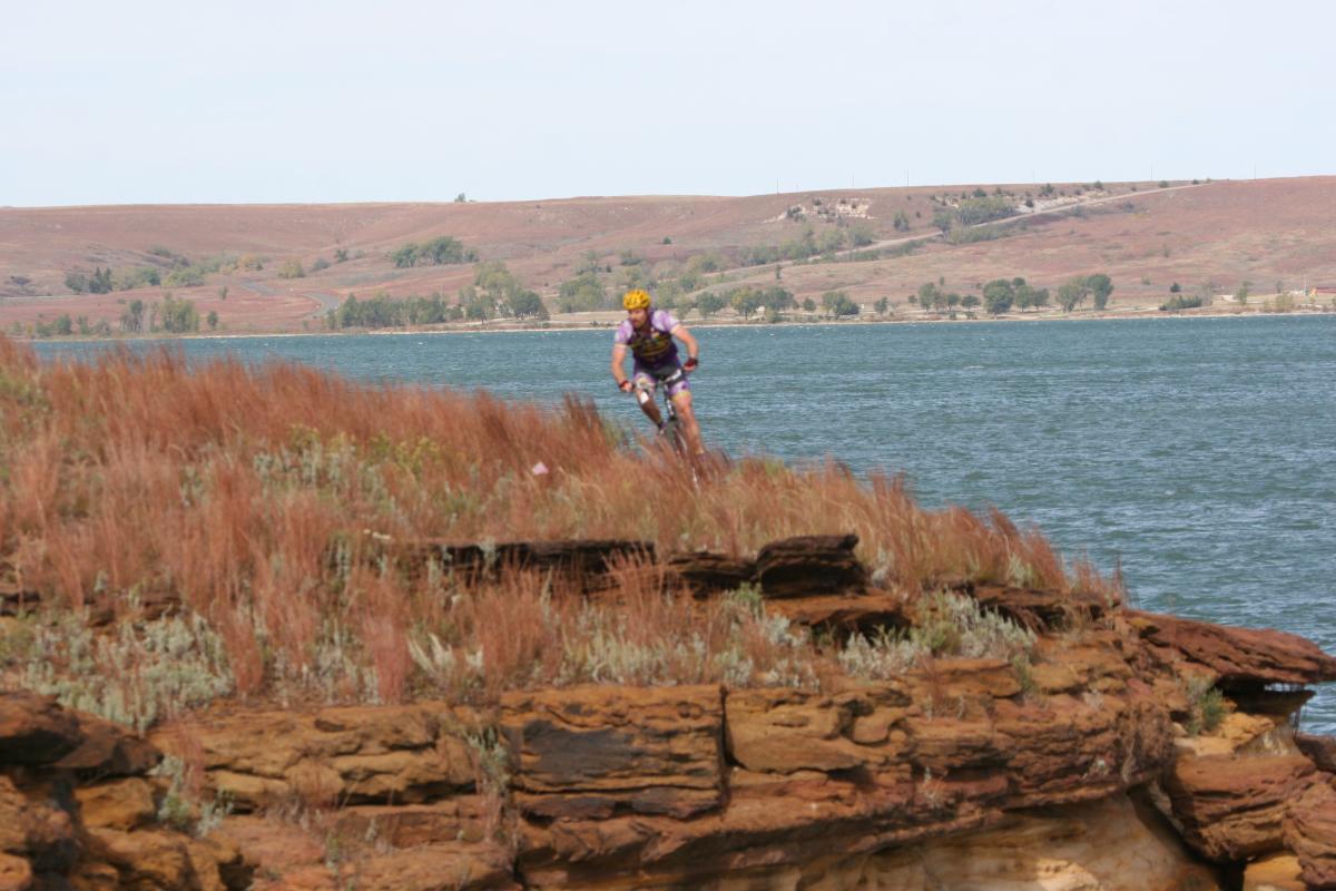 A mountain biker navigates the edge of a rocky cliff with tall grasses in the foreground, overlooking a body of water. In the background, green hills and trees line the shore, with a clear blue sky above. Switchgrass mountain bike trail.