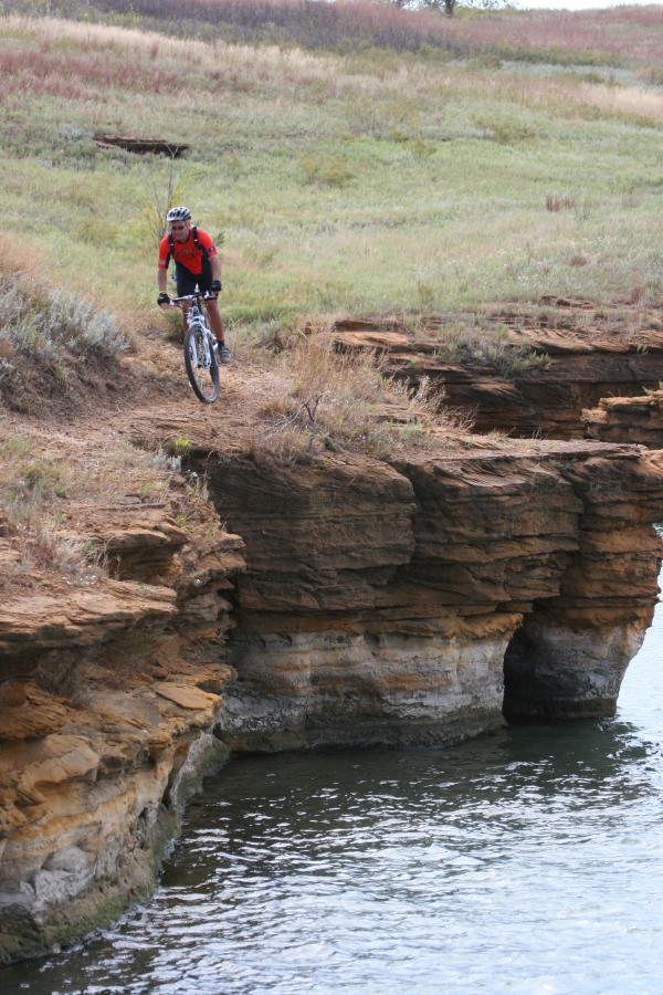 A mountain biker wearing a red shirt and helmet is riding along the edge of a rocky cliff, overlooking a body of water. The landscape features green grass and rocky formations, with a gentle slope leading down to the water's surface. Switchgrass mountain bike trail.