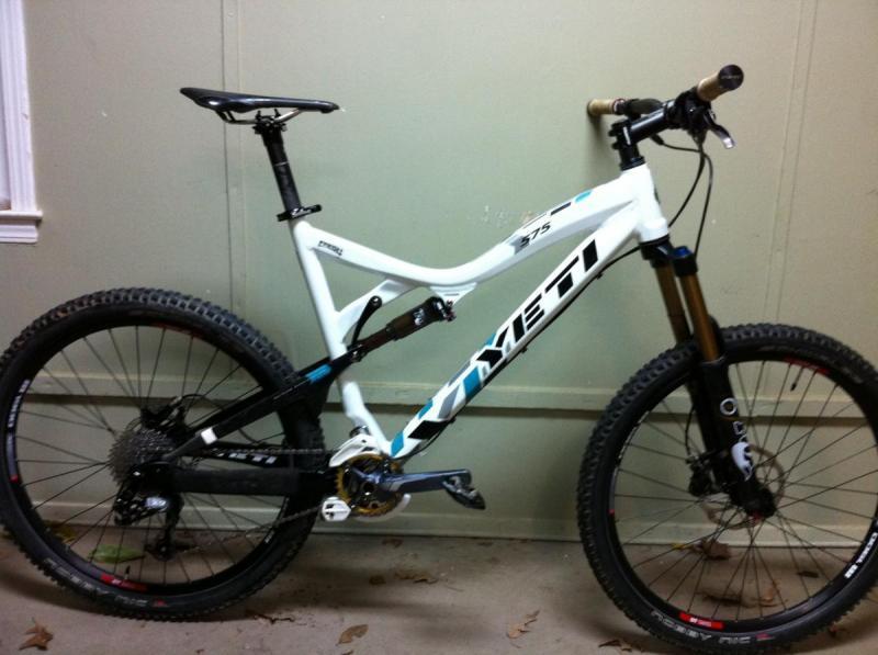Yeti 575: Alt text: A white Yeti mountain bike with blue and black accents, showing front and rear wheels, suspension, and handlebars, positioned against a light green wall.