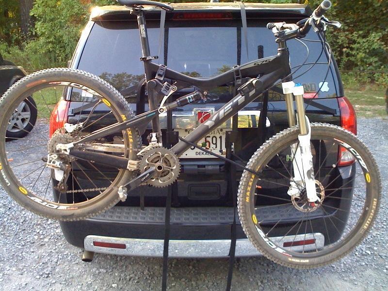 Yeti 575: A black mountain bike mounted on the back of an SUV, with its front wheel suspended in the air. The bike features a dual suspension system and is secured with straps. The vehicle is parked on gravel, surrounded by greenery in the background.