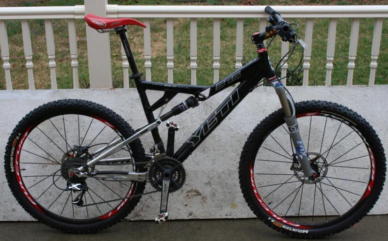 Yeti 575: Alt text: A side view of a black and silver mountain bike with prominent red accents on the seat and wheel rims, resting on a porch. The bike features wide tires, front suspension, and multiple gears.