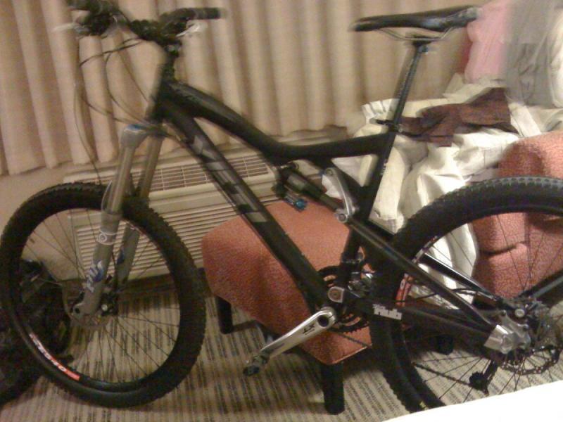 Yeti 575: A black and silver mountain bike leaning against an armchair in a hotel room, with a bed and window curtains visible in the background.