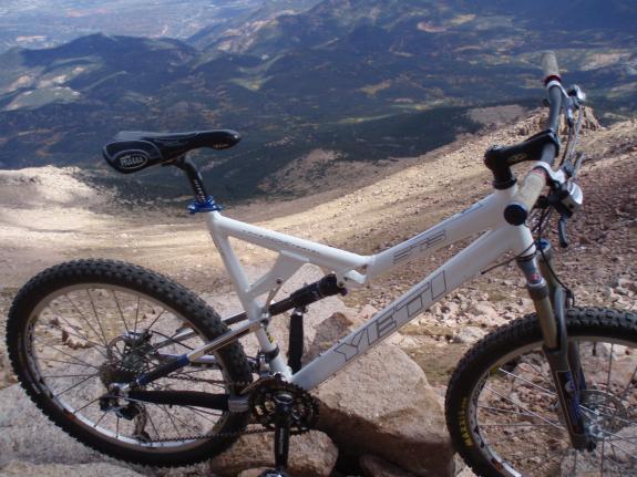 Yeti 575: A white mountain bike is parked on rocky terrain, overlooking a vast landscape of mountains and valleys below. The bike's frame features a bold logo, and its tires are suited for rugged trails. The scene captures a clear day with a panoramic view of the surroundings.