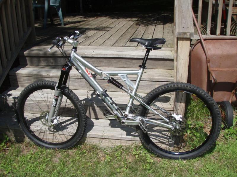 Yeti 575: A silver mountain bike with a sturdy frame and thick tires, leaning against a wooden porch. The bike features a suspension system and is positioned on green grass, with a wooden deck and a wheelbarrow visible in the background.
