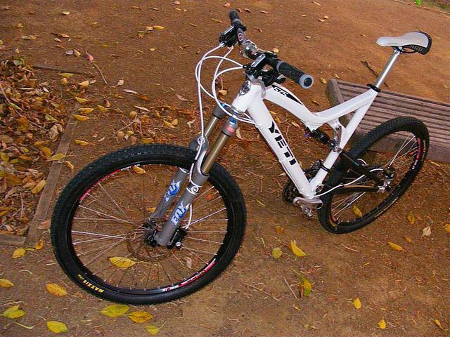 Yeti 575: A white mountain bike with a shiny fork and black tires, resting on a bed of fallen leaves in a dirt area. The bike features a prominent brand logo on the frame and has a sleek, modern design.