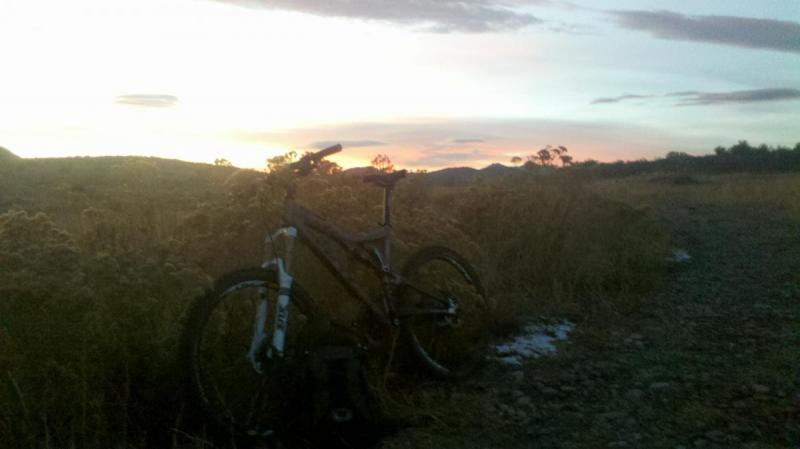 Yeti 575: A mountain bike stands beside a gravel path, surrounded by tall grass and wildflowers, with a colorful sunset illuminating the sky in shades of orange and purple. Silhouetted hills are visible in the background, creating a serene and tranquil outdoor scene.
