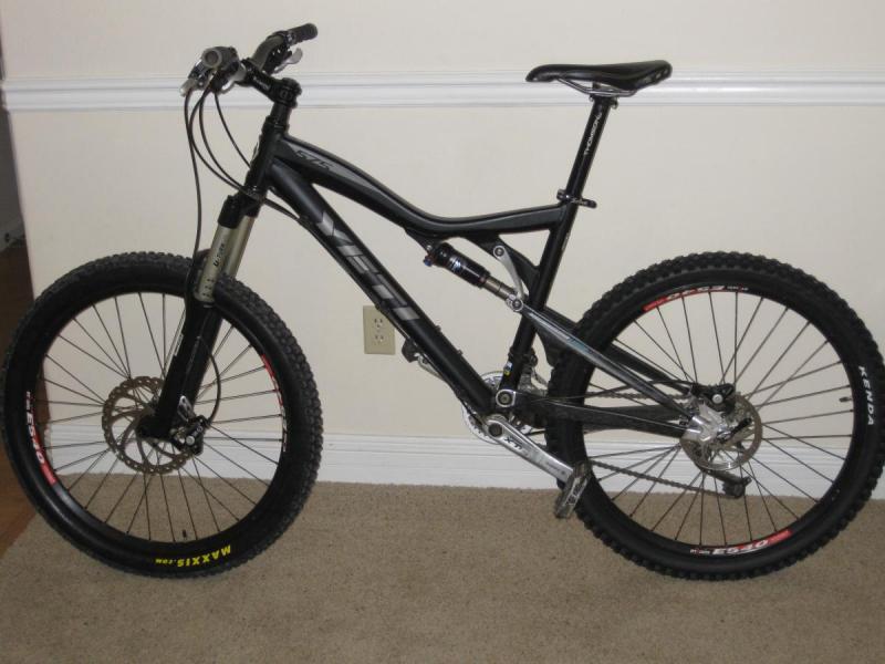 Yeti 575: A black mountain bike with a prominent frame, featuring wide tires and adjustable suspension. The bike is positioned against a neutral-colored wall with a carpeted floor. The brand name "Yeti" is visible on the frame, and it has disc brakes on both wheels.
