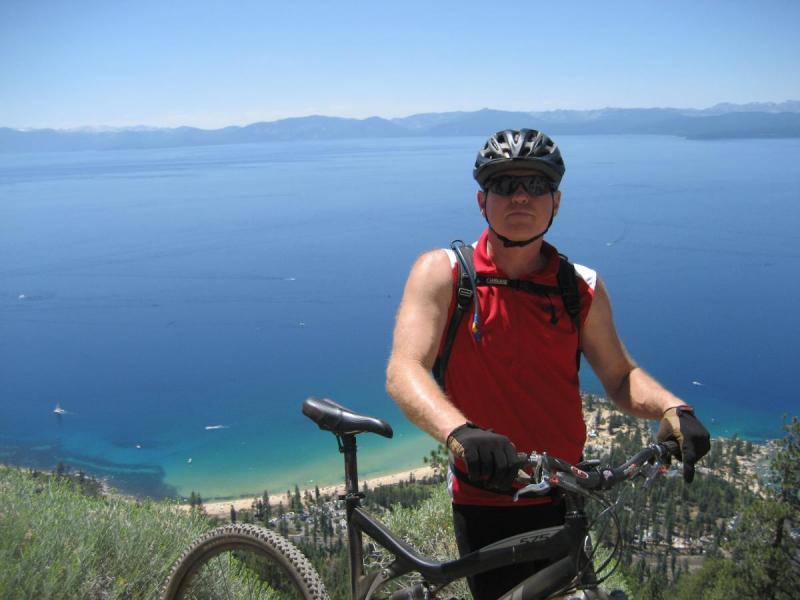Yeti 575: A person wearing a red sleeveless shirt, sunglasses, and a helmet stands next to a mountain bike, overlooking a scenic view of a lake and coastline under a clear blue sky. The background features mountains in the distance and a lush green landscape.