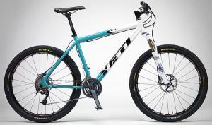 Yeti ARC: Mountain bike by Yeti, featuring a turquoise and white color scheme, front suspension, and wide tires suitable for rugged terrain. The bike is displayed against a gray background, showcasing its sleek design and components.