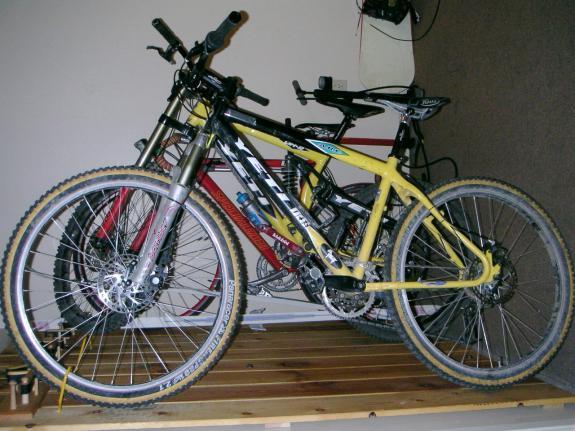 Yeti ARC: A close-up image of two mountain bikes with distinctive colors. The primary bike in the foreground is yellow and black, featuring thick tires and a sporty design. The background includes a red mountain bike partially visible. Both bikes are displayed on a wooden platform in an indoor setting.