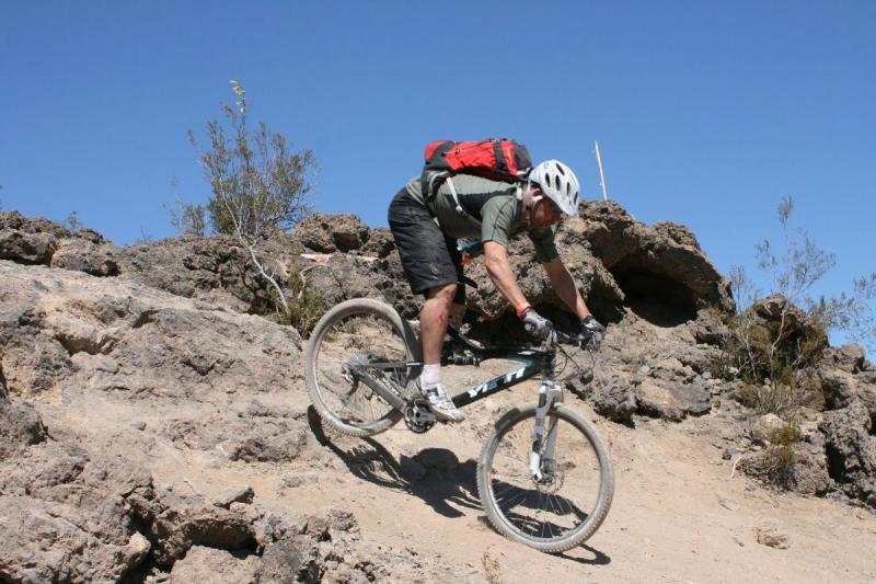 Yeti ASR 5 Alloy: A mountain biker navigating a rocky terrain on a bright, clear day. The cyclist is wearing a helmet, gloves, and a backpack, leaning forward as they descend the uneven path surrounded by rocks and sparse vegetation.
