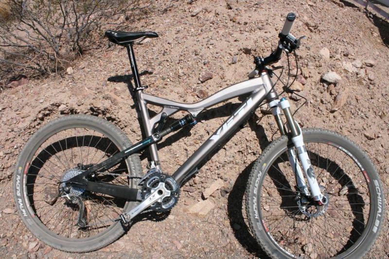 Yeti ASR 5 Alloy: A mountain bike is positioned on rocky terrain, showcasing its sleek frame and suspension system. The bike features large tires designed for rugged surfaces, with visible components like the gears and handlebars. In the background, sparse vegetation is present, indicating a natural outdoor setting.